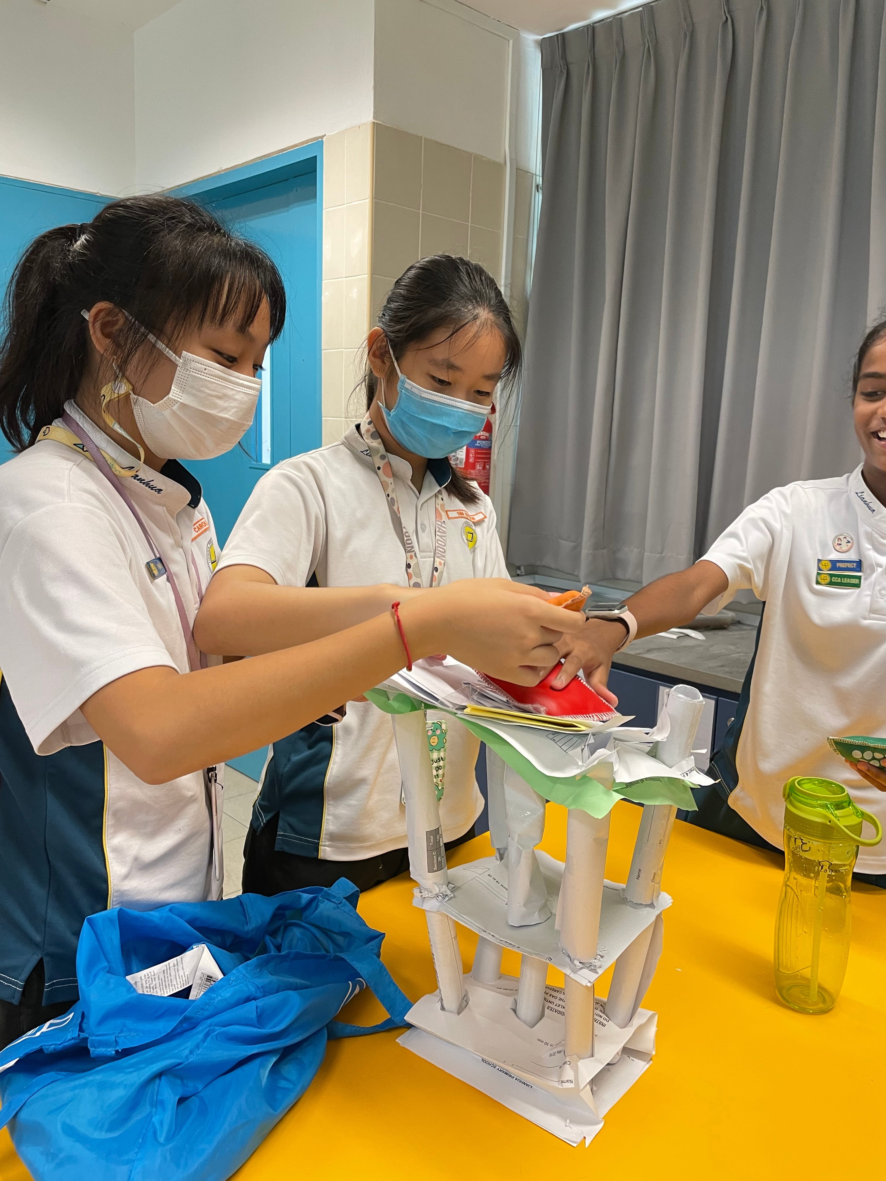Three masked students in white uniforms work together building a paper tower structure on a yellow table.